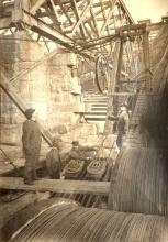 Stringing cable on the Williamsburg Bridge. Othniel Nichols and Leffert Buck in work clothes looking up as cable is strung. Two other men are visible near the cable shoes.
